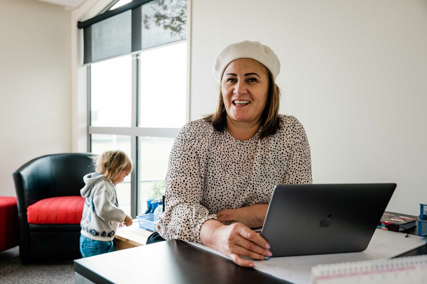 Woman sits at a desk behind a laptop, looking at the camera with her mokopuna in the background.